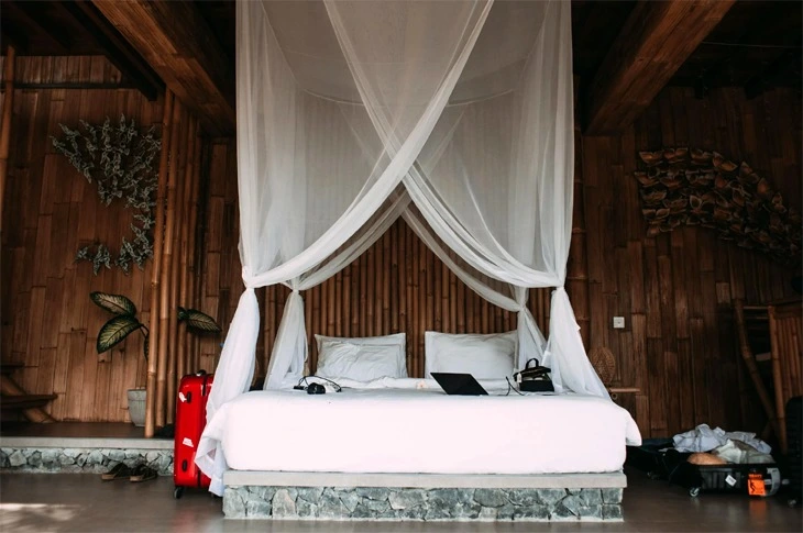 A canopy bed frame with an elegant, four-poster design, made of dark wood, and draped with sheer white curtains, surrounding a cozy, well-made bed.